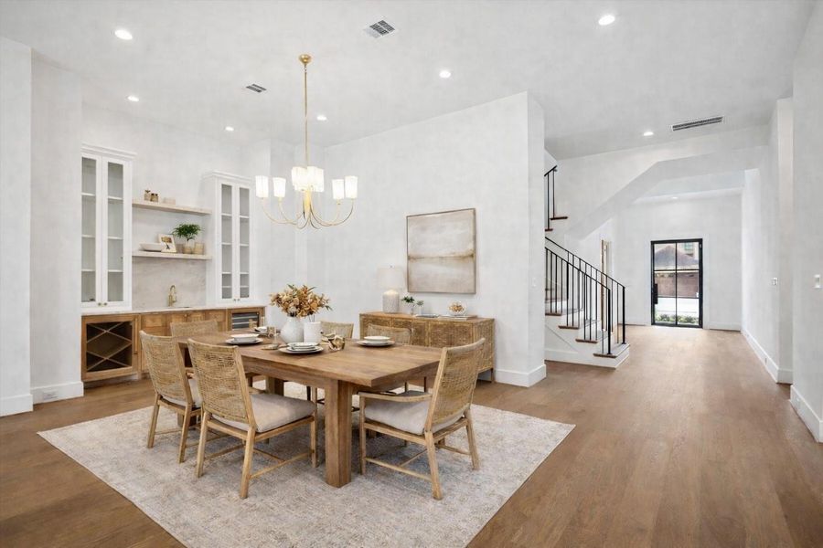 Formal dining space centered beneath a statement brass chandelier, staged with a solid wood table and woven accent chairs for a warm, designer feel. Custom built in dry bar with wine storage, glass front cabinetry, and stone backsplash adds entertaining appeal, while wide plank hardwood floors and the open staircase create seamless flow from dining to entry. *Virtually Staged*