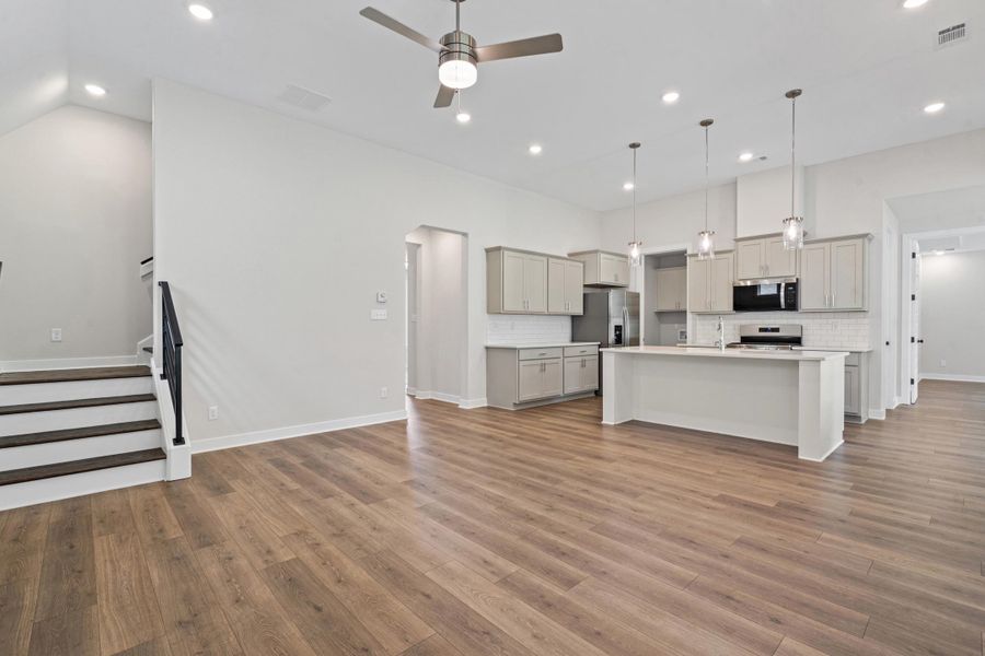 Spacious open-concept living area featuring wood-finish flooring, a contemporary kitchen island with pendant lighting, recessed ceiling lights, a ceiling fan, and a staircase with dark wood treads and a black handrail Spacious open-concept living area featuring wood-finish flooring, a contemporary kitchen island with pendant lighting, recessed ceiling lights, a ceiling fan, and a staircase with dark wood treads and a black handrail