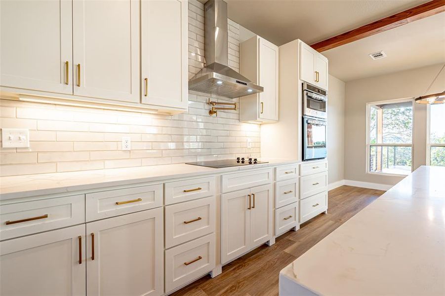 Kitchen with wall chimney range hood, light stone counters, white cabinetry, tasteful backsplash, and beam ceiling Kitchen with wall chimney range hood, light stone counters, white cabinetry, tasteful backsplash, and beam ceiling