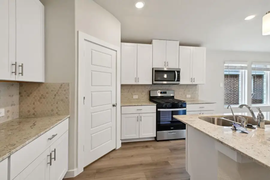 This modern kitchen features sleek white shaker cabinets, stainless steel appliances, and a central island with a double sink. The light countertops and octagon backsplash complement the natural light from the large windows, creating a bright and inviting space.