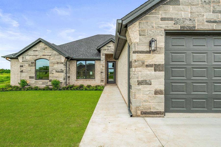 Entrance to property featuring stone siding, an attached garage, a lawn, and a shingled roof