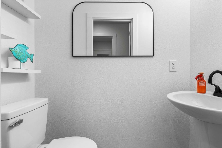 This is a modern, compact bathroom featuring a pedestal sink and a simple toilet. It has a wall-mounted mirror and open shelving with decorative accents. The space is bright with a clean, minimalist design.