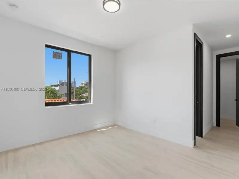 Neutral interior featuring light-toned wood-finish flooring, crisp white walls, a multi-pane window with dark trim, and a ceiling-mounted flush-mount light fixture Neutral interior featuring light-toned wood-finish flooring, crisp white walls, a multi-pane window with dark trim, and a ceiling-mounted flush-mount light fixture