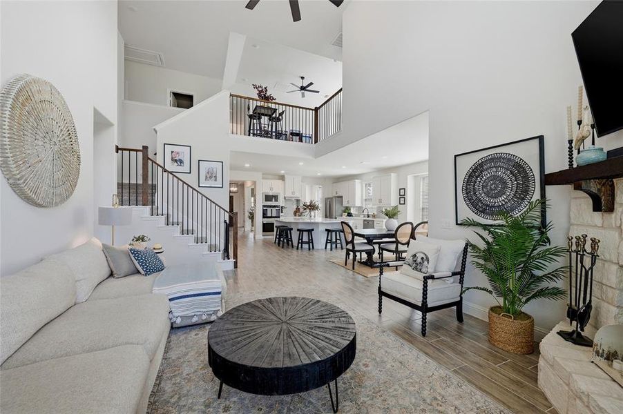 Open-concept living space featuring high ceilings, a two-story staircase with dark wood banisters and black iron spindles, wood-finish flooring throughout, a stone-clad fireplace with a dark wood mantel, and a kitchen with a large island and white cabinetry