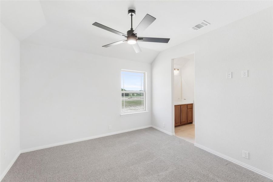 Empty bedroom with vaulted ceiling, ceiling fan, carpet flooring, and ensuite bathroom doorway visible. Empty bedroom with vaulted ceiling, ceiling fan, carpet flooring, and ensuite bathroom doorway visible.