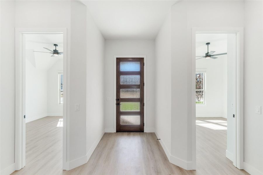 Hallway with wood and glass front door, light hardwood floors, and open doorways to rooms with ceiling fans