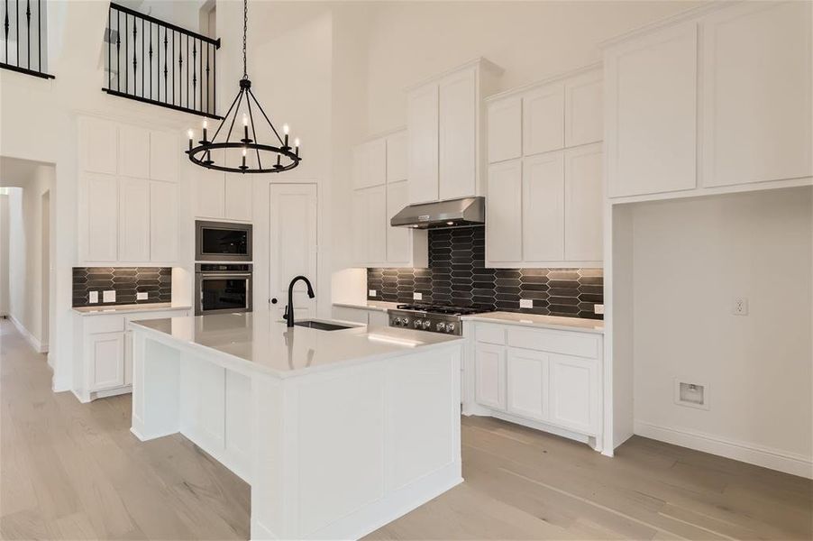 Kitchen featuring an island with sink, stainless steel appliances, wall chimney range hood, sink, and white cabinets