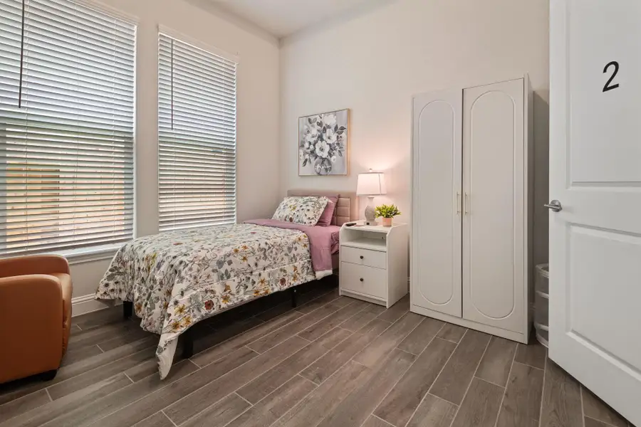 Cozy bedroom with natural light, featuring a single bed with floral bedding, a nightstand with a lamp, a wardrobe, and wood-look tile flooring. Cozy bedroom with natural light, featuring a single bed with floral bedding, a nightstand with a lamp, a wardrobe, and wood-look tile flooring.