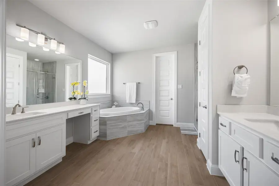 The primary bath features separated dual vanities, wood tiled floors, a spacious walk-in shower, and a luxurious soaking tub—designed for both function and relaxation.