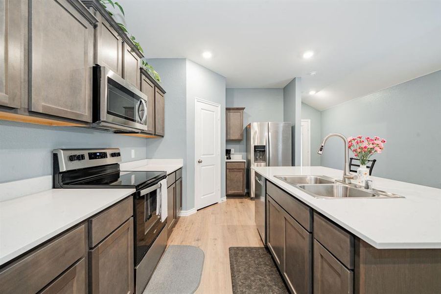 Kitchen with appliances with stainless steel finishes, a kitchen island with sink, light wood-type flooring, dark brown cabinets, and vaulted ceiling Kitchen with appliances with stainless steel finishes, a kitchen island with sink, light wood-type flooring, dark brown cabinets, and vaulted ceiling