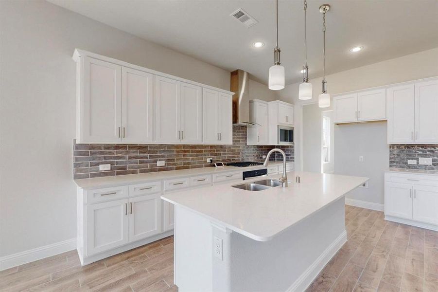 Kitchen featuring wood tiled floors, decorative backsplash, hanging light fixtures, a center island with sink, and white cabinetry