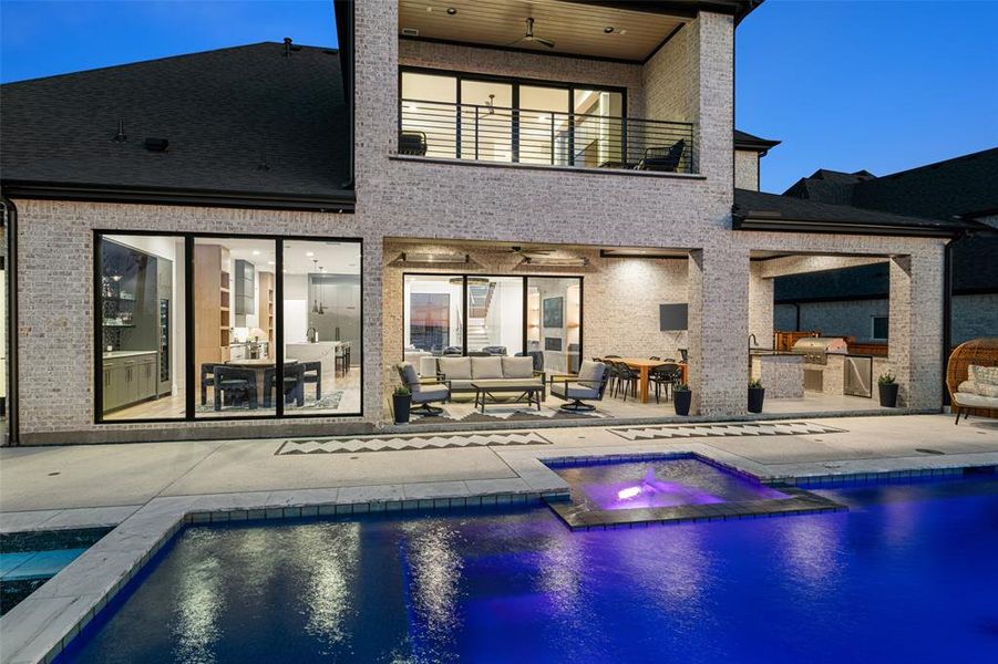 Resort-style backyard retreat featuring a stunning pool and spa with ambient lighting, expansive patio space, and multiple covered outdoor living areas. The outdoor kitchen with built-in grill and bar seating creates the perfect setting for entertaining, while the upper balcony and walls of glass showcase the home’s seamless indoor-outdoor design and views of the beautifully illuminated outdoor space.