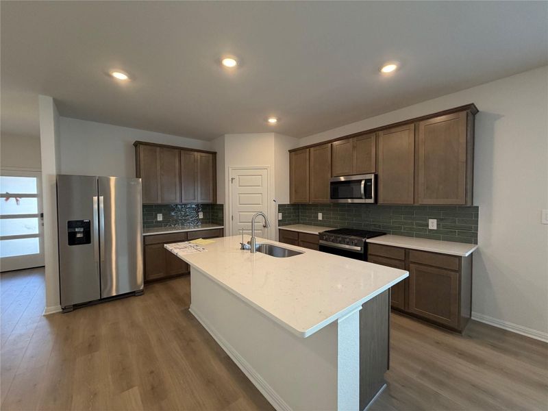Kitchen featuring stainless steel appliances, dark brown cabinetry, light stone counters, a center island with sink, and dark wood-style floors
