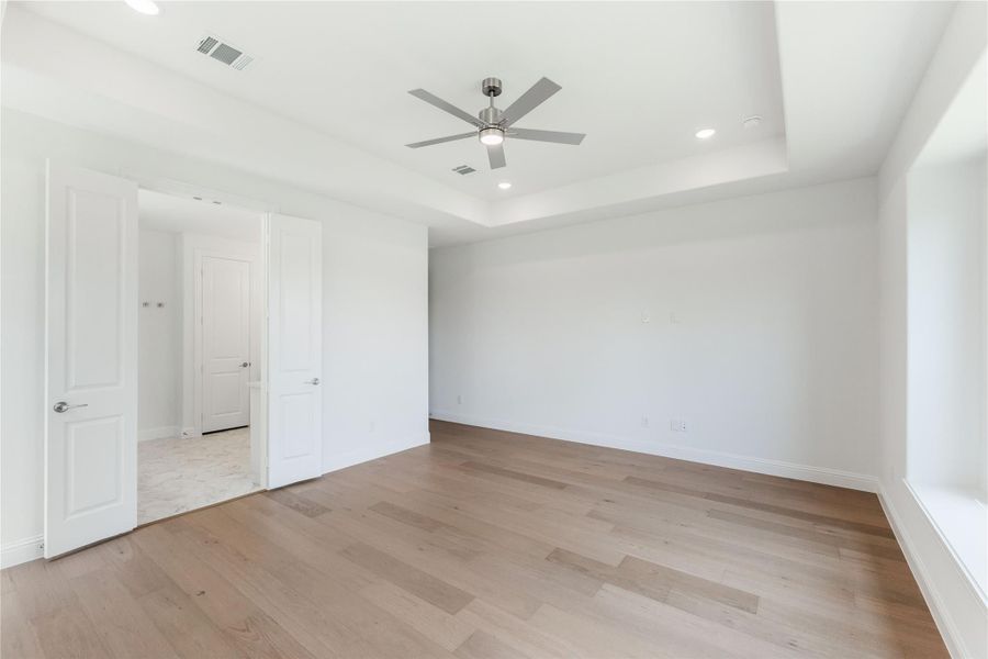 Empty bedroom with tray ceiling, ceiling fan, hardwood floors, and open door to bathroom