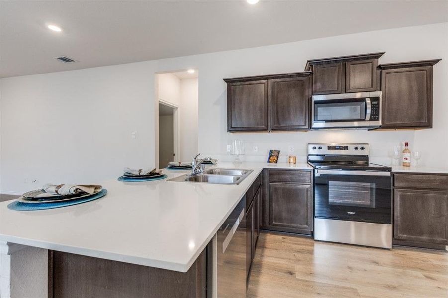 Kitchen with stainless steel appliances, dark brown cabinetry, light wood-style flooring, recessed lighting, and light stone countertops Kitchen with stainless steel appliances, dark brown cabinetry, light wood-style flooring, recessed lighting, and light stone countertops