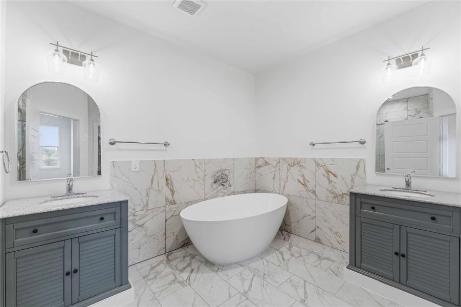 Luxurious primary bath featuring a stunning freestanding soaking tub framed by elegant marble-style tile. Dual vanities with custom cabinetry, arched mirrors, and modern lighting create a spa-like atmosphere. Bright, clean finishes and thoughtful design offer both beauty and comfort.