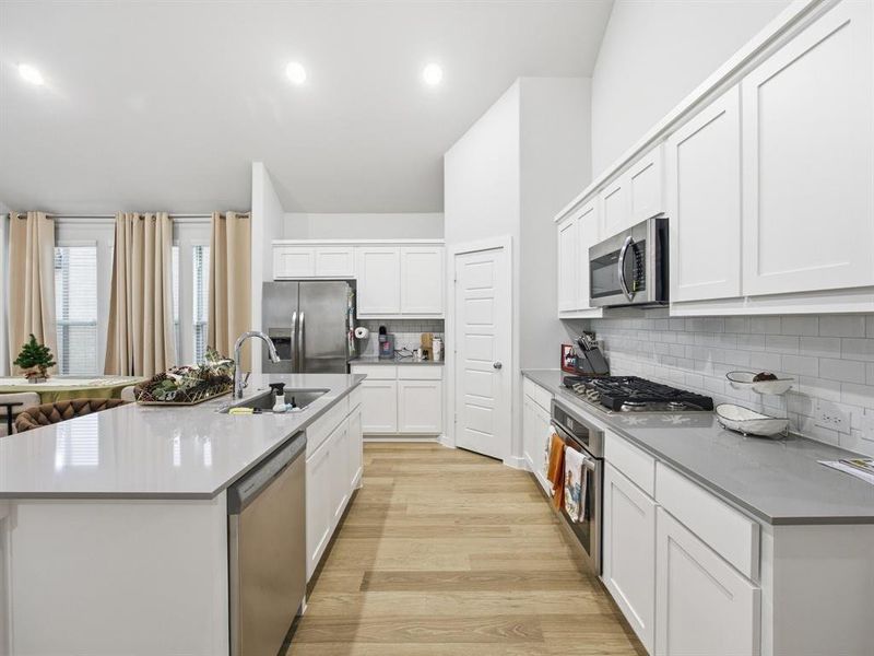 Kitchen featuring appliances with stainless steel finishes, light wood-style flooring, an island with sink, backsplash, and white cabinetry