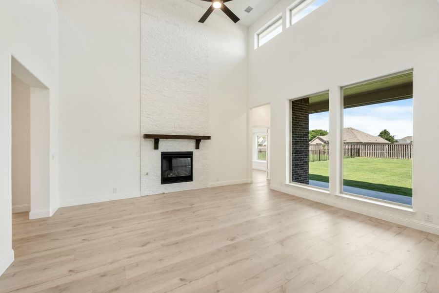 Open living room with high ceilings, stone fireplace, light wood floors, and large windows overlooking backyard