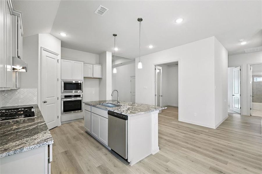 Kitchen with stainless steel appliances, light wood-type flooring, light stone counters, decorative backsplash, and a kitchen island with sink Kitchen with stainless steel appliances, light wood-type flooring, light stone counters, decorative backsplash, and a kitchen island with sink