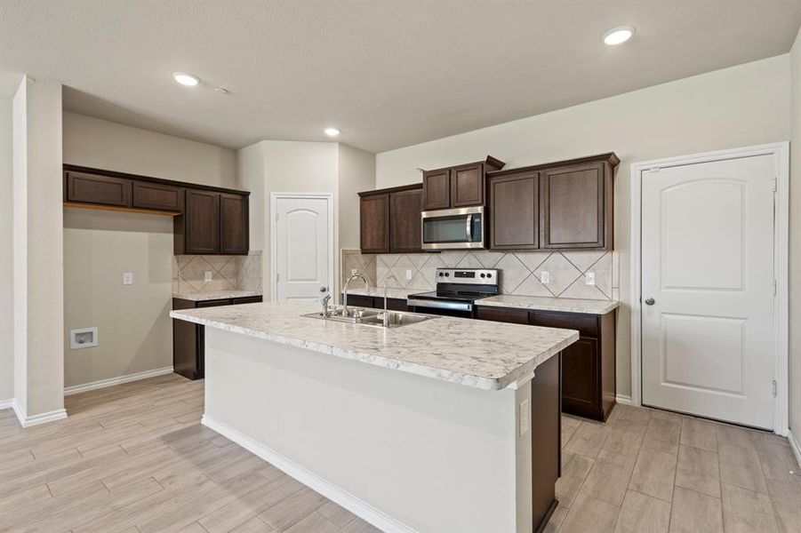 Kitchen featuring backsplash, light countertops, light wood-style floors, a center island with sink, and dark brown cabinetry Kitchen featuring backsplash, light countertops, light wood-style floors, a center island with sink, and dark brown cabinetry