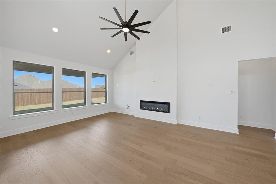 Unfurnished living room with high vaulted ceiling, a glass covered fireplace, light wood-style floors, a ceiling fan, and recessed lighting Unfurnished living room with high vaulted ceiling, a glass covered fireplace, light wood-style floors, a ceiling fan, and recessed lighting