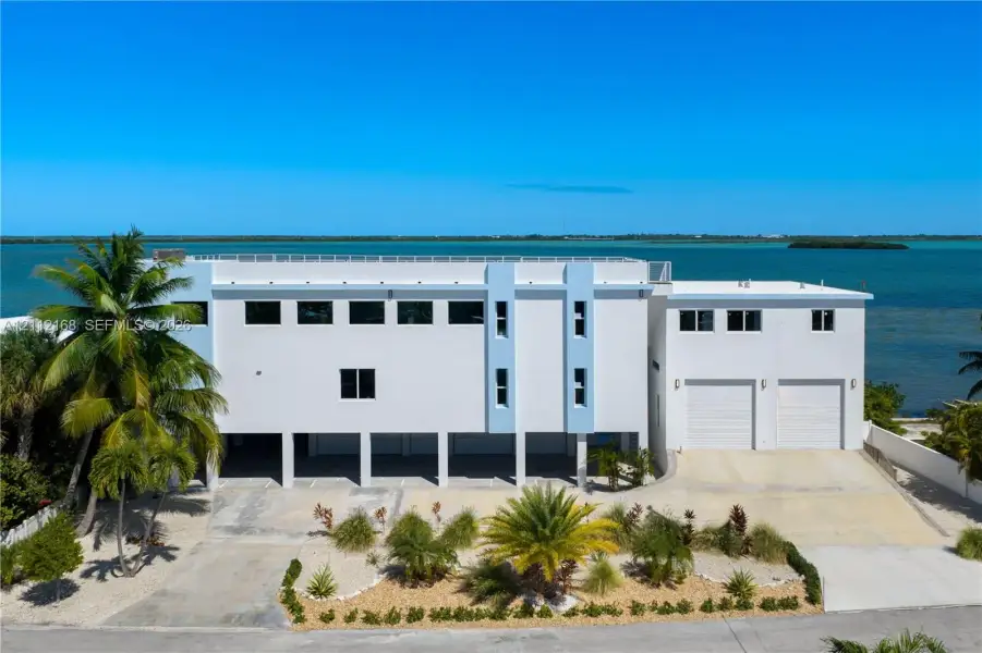Multi-level contemporary exterior featuring a concrete facade, two-car garage, covered ground-level parking, rooftop railing, and waterfront orientation Multi-level contemporary exterior featuring a concrete facade, two-car garage, covered ground-level parking, rooftop railing, and waterfront orientation