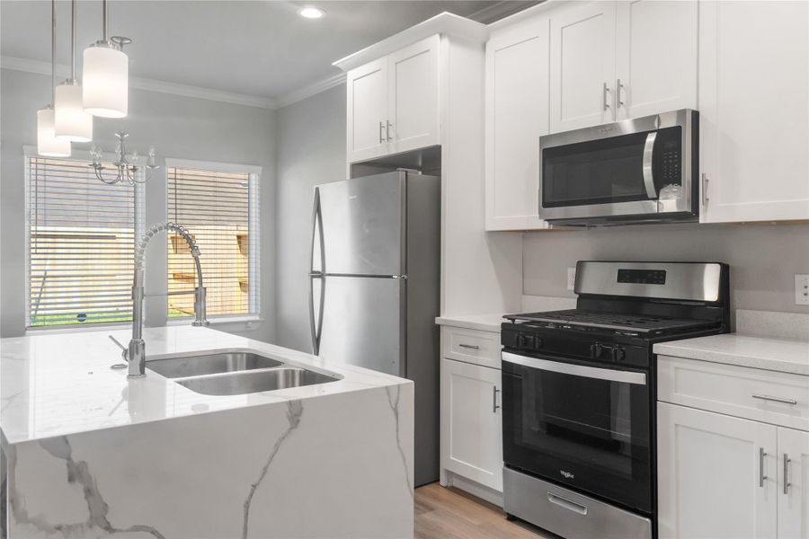 This modern kitchen features sleek white cabinets, stainless steel appliances, and a stylish marble-look island with a dual sink. Bright and inviting, it also includes pendant lighting and large windows for natural light.