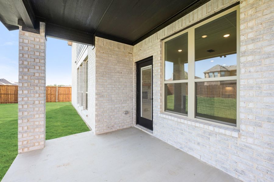 Covered back patio with concrete floor, light brick exterior, black door, and fenced backyard with green lawn.