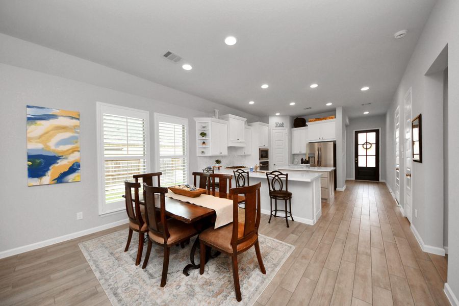 This photo showcases a modern, open-concept dining and kitchen area with sleek wood flooring. The dining space features a wooden table and chairs set on a stylish rug, while the kitchen boasts white cabinetry, a large island with bar seating, and stainless steel appliances. Ample natural light streams in through large windows, creating a bright and welcoming atmosphere.