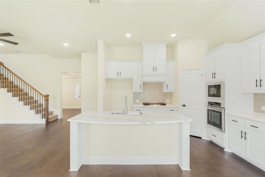 Open kitchen with white cabinets, large island, built-in ovens, and dark hardwood floors, open to staircase area. Open kitchen with white cabinets, large island, built-in ovens, and dark hardwood floors, open to staircase area.