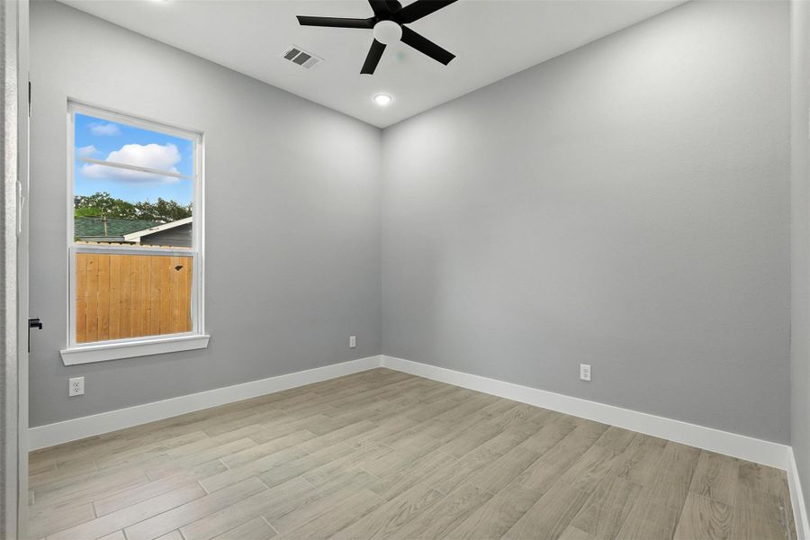 This room features a modern, minimalist design with light gray walls and wood-look flooring. It has a large window offering natural light, a ceiling fan, and recessed lighting, creating a bright and airy atmosphere.