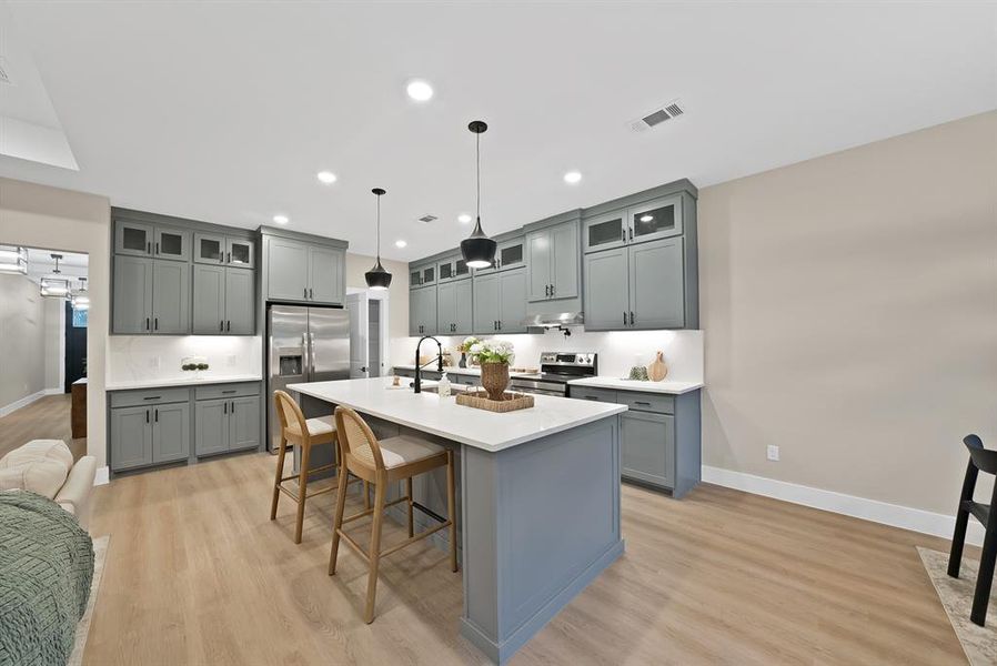 Open-concept kitchen featuring an expansive center island with seating, grey shaker-style cabinetry, stainless steel appliances, white countertops, and wood-finish flooring Open-concept kitchen featuring an expansive center island with seating, grey shaker-style cabinetry, stainless steel appliances, white countertops, and wood-finish flooring