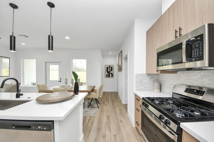 This photo showcases a modern kitchen with sleek appliances, including a gas stove and microwave. The space features a bright, open layout with white countertops and wood cabinets, leading to a cozy dining area with natural light from large windows. Contemporary lighting fixtures and stylish decor complete the look. This photo showcases a modern kitchen with sleek appliances, including a gas stove and microwave. The space features a bright, open layout with white countertops and wood cabinets, leading to a cozy dining area with natural light from large windows. Contemporary lighting fixtures and stylish decor complete the look.