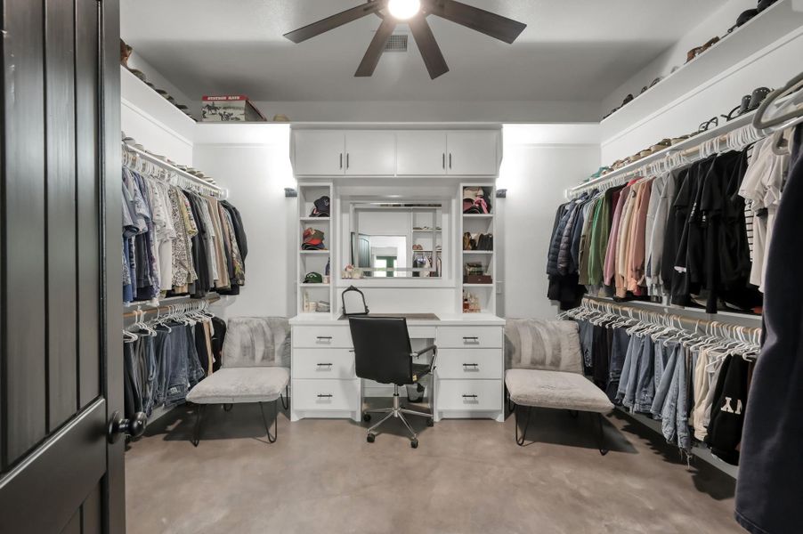 This spacious walk-in closet features ample hanging space, built-in shelving, and a vanity with seating. It includes modern lighting and a ceiling fan, providing a functional and stylish dressing area.