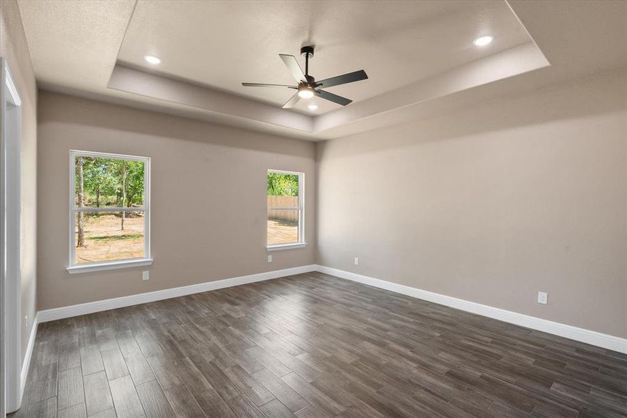 Primary bedroom featuring tray ceiling with recessed lighting and a ceiling fan, wood-finish flooring, two windows, and neutral wall tones. Individual bedroom remote for integrated lighting.