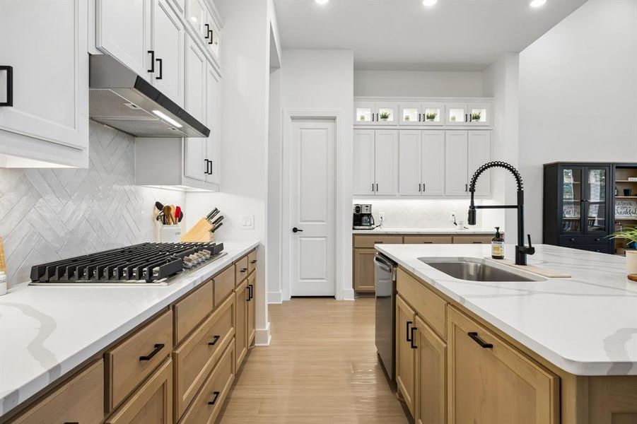 Gourmet kitchen featuring a gas cooktop, herringbone tile backsplash, white and wood-finish cabinetry, light-toned flooring, and an island with an undermount sink and gooseneck faucet Gourmet kitchen featuring a gas cooktop, herringbone tile backsplash, white and wood-finish cabinetry, light-toned flooring, and an island with an undermount sink and gooseneck faucet