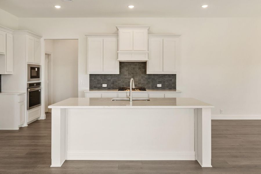 Kitchen with white cabinets, large island with sink, gray tile backsplash, and stainless steel double ovens on hardwood floors Kitchen with white cabinets, large island with sink, gray tile backsplash, and stainless steel double ovens on hardwood floors