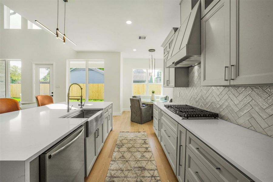 This modern kitchen features sleek gray cabinetry, a stylish herringbone backsplash, and a large island with a built-in sink and dishwasher. It boasts ample natural light from large windows and is accented by contemporary lighting fixtures.