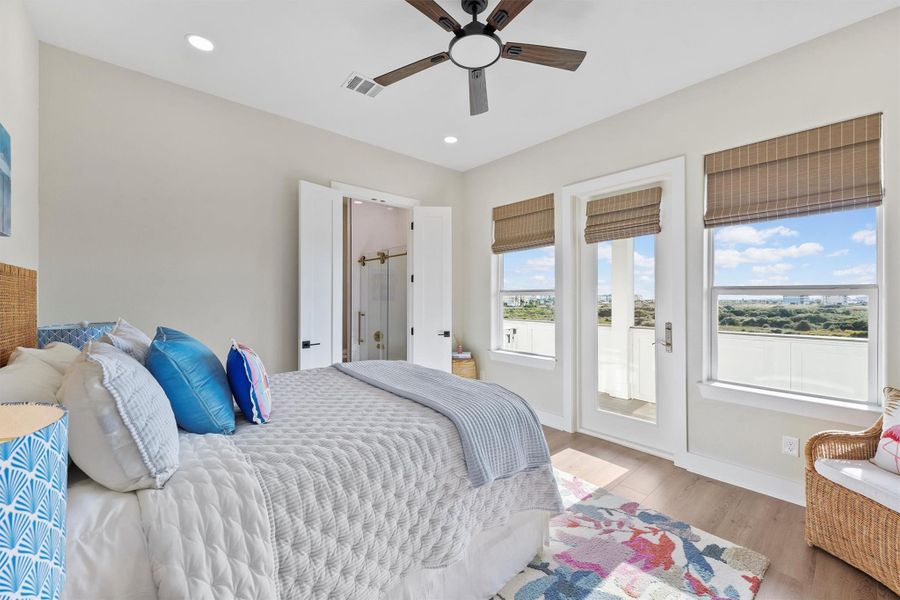 This secondary Queen room, also located on the second floor, is a bright bedroom featuring a queen bed with colorful pillows, a ceiling fan, and access to a balcony through hurricane rated glass door. Large windows offer natural light and scenic views, while an en-suite bathroom is visible through open double doors with a walk in closet