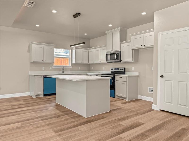 Kitchen featuring a central island with a solid surface countertop, white cabinetry with black hardware, wood-finish flooring, recessed lighting, and a stainless steel over-the-range microwave Kitchen featuring a central island with a solid surface countertop, white cabinetry with black hardware, wood-finish flooring, recessed lighting, and a stainless steel over-the-range microwave