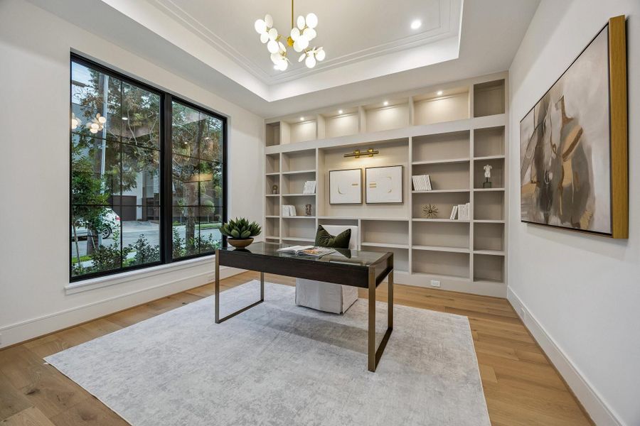 Off the entrance of the home is a sophisticated study designed with warmth, elegance, and functionality in mind. The focal point of the room is the expansive built-in shelving wall finished in a muted warm tone. The lighting design is layered and elegant. To the left, large black-framed windows stretch nearly floor-to-ceiling, allowing natural light to filter gently into the room while maintaining an airy feel. The tray ceiling adds a more polished, expansive and luxurious presence.