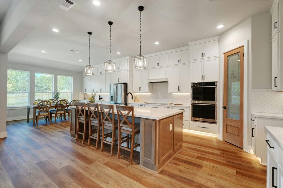 Spacious kitchen featuring an expansive center island with seating, wood-finish flooring, white cabinetry, double wall ovens, and a pantry with a frosted glass door