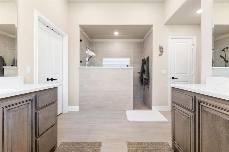 Spacious bathroom featuring a walk-in shower with light-toned tile, dual vanity cabinets with stone countertops, and recessed lighting Spacious bathroom featuring a walk-in shower with light-toned tile, dual vanity cabinets with stone countertops, and recessed lighting