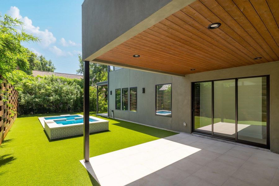 Expansive outdoor living area featuring a covered patio with wood-finish ceiling and recessed lighting, a modern pool, artificial turf, and a privacy fence