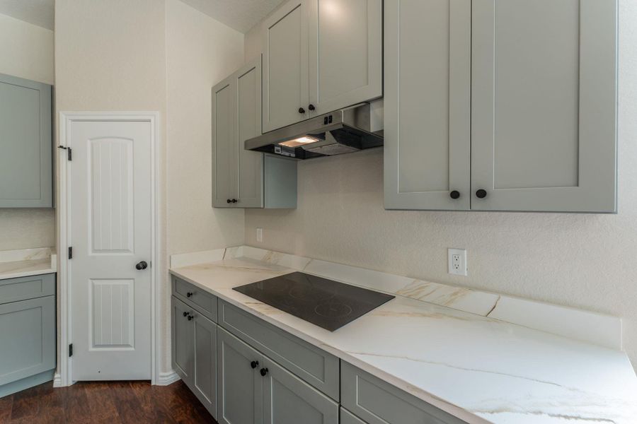 Modern kitchen featuring light gray cabinetry with black hardware, white countertops with subtle veining, an integrated black electric cooktop, and a stainless steel range hood