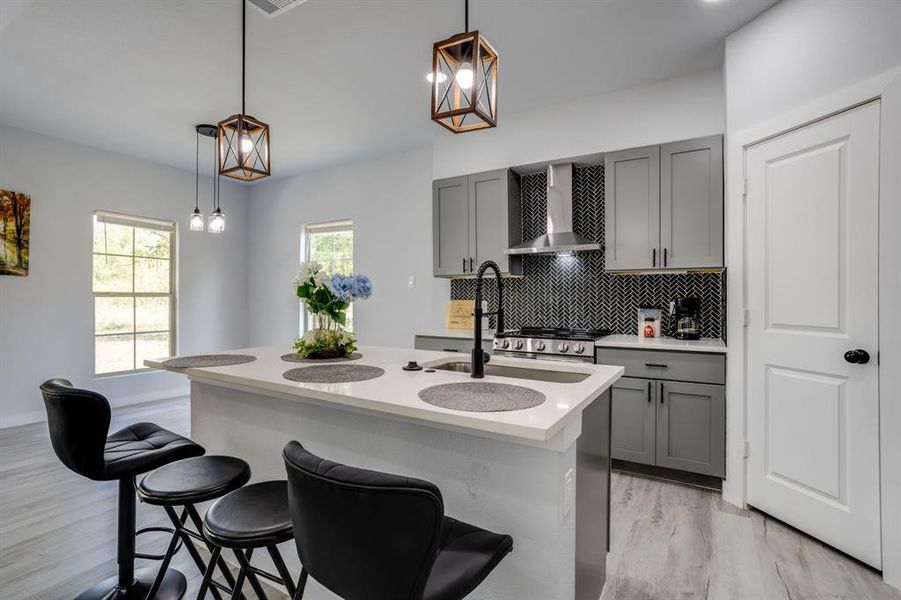 Kitchen with gray cabinets, backsplash, decorative light fixtures, and wall chimney range hood Kitchen with gray cabinets, backsplash, decorative light fixtures, and wall chimney range hood