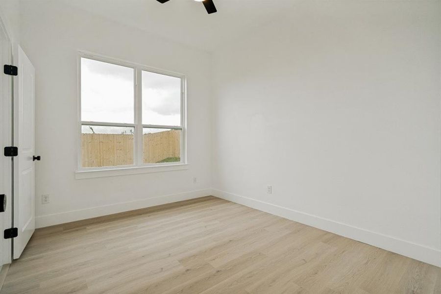 Bright interior featuring wood-finish flooring, a large window, a white door with black hardware, white walls, and a ceiling fan Bright interior featuring wood-finish flooring, a large window, a white door with black hardware, white walls, and a ceiling fan