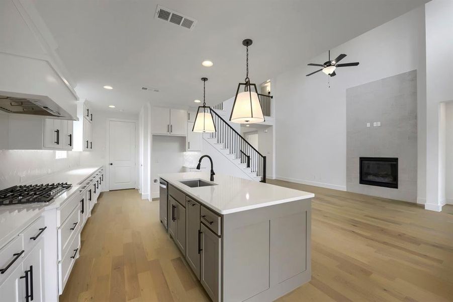 Open-concept living area featuring a kitchen island with pendant lighting, a gas range, wood-finish flooring, a fireplace with a textured surround, and a staircase with a black metal railing Open-concept living area featuring a kitchen island with pendant lighting, a gas range, wood-finish flooring, a fireplace with a textured surround, and a staircase with a black metal railing