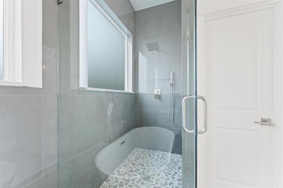 This modern bathroom features a sleek walk-in shower with glass doors, a rainfall showerhead, and a stylish freestanding tub. The space is brightened by a large frosted window and elegant gray tile walls.