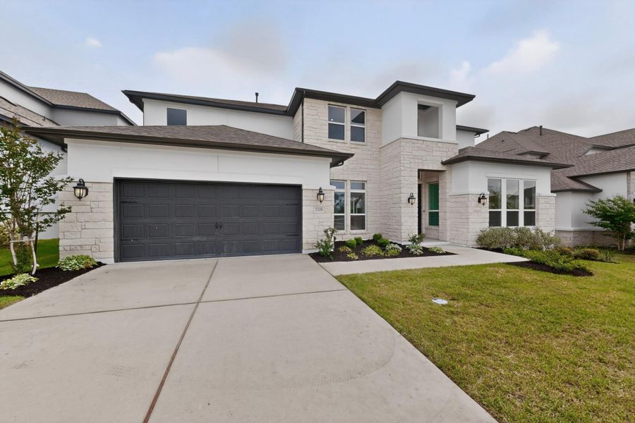 Contemporary two-story residence featuring a stone and stucco exterior, an attached two-car garage with a dark panel door, a concrete driveway, and front landscaping